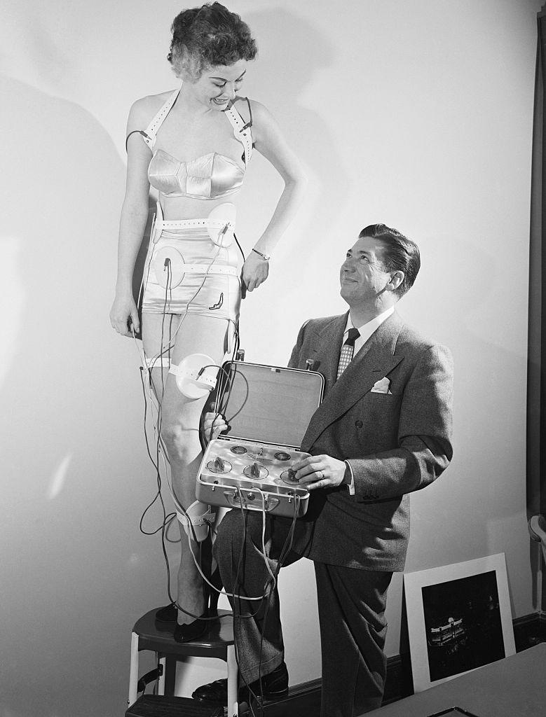 Burton Skiles, president of Relaxacizor Co., turns on the Relaxacizor's current that will give model Shirley Saxby the 'reducing-while-relaxing' treatment for a streamlined figure. Chicago, Illinois, 1950.jpg