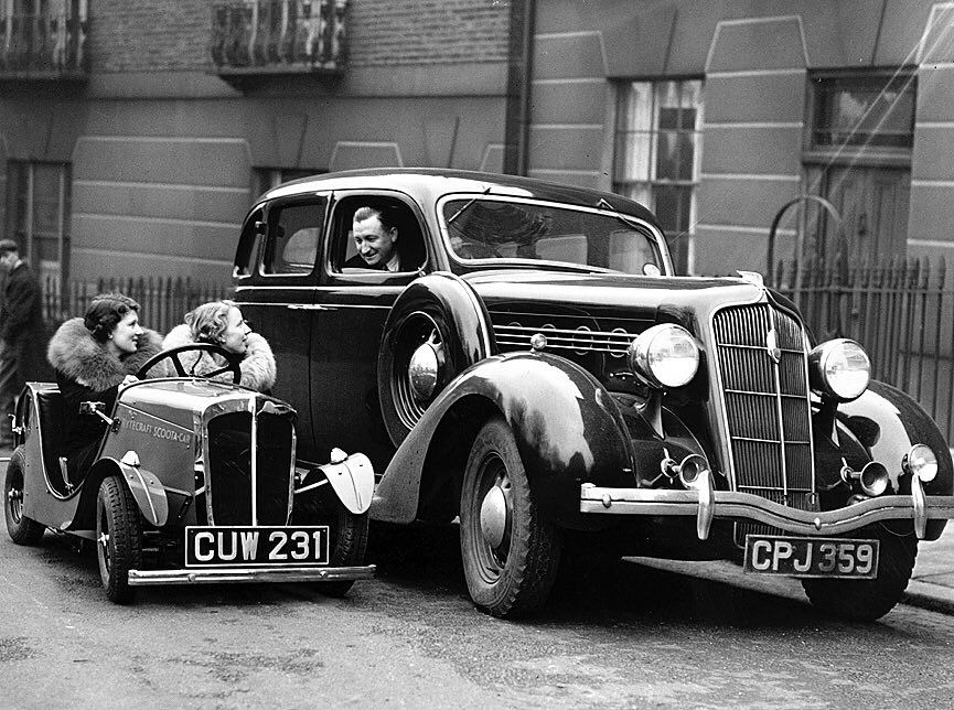 A newly-designed 2.5 horse power Rytecraft Scootacar, during a demonstration in London. The little car, which can carry two people at speeds up to 15mph, is dwarfed by an ordinary-sized vehicle (1936).jpg
