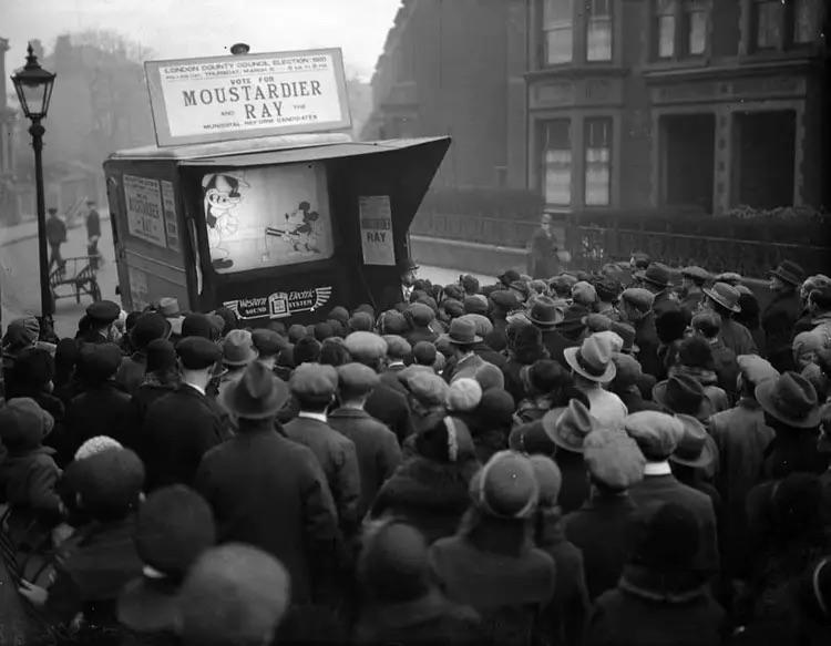 A crowd gathers to watch Mickey Mouse on a small screen on a London street, 1931.jpg