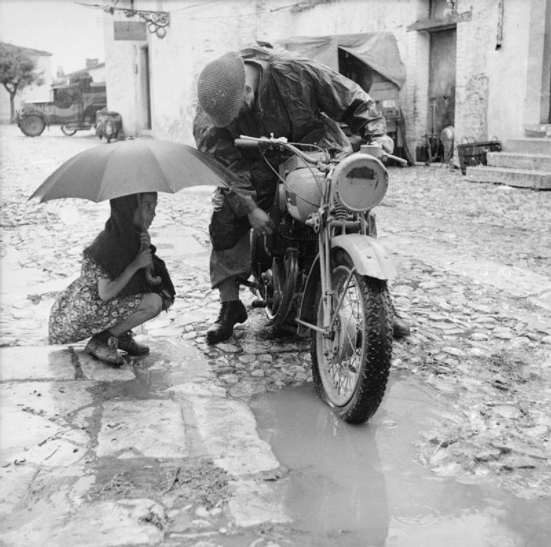 80 years ago, a little girl holding an umbrella watches a British despatch rider attempt to clear the carbuerettor of his motorcycle in torrential rain, somewhere in southern Italy.jpg