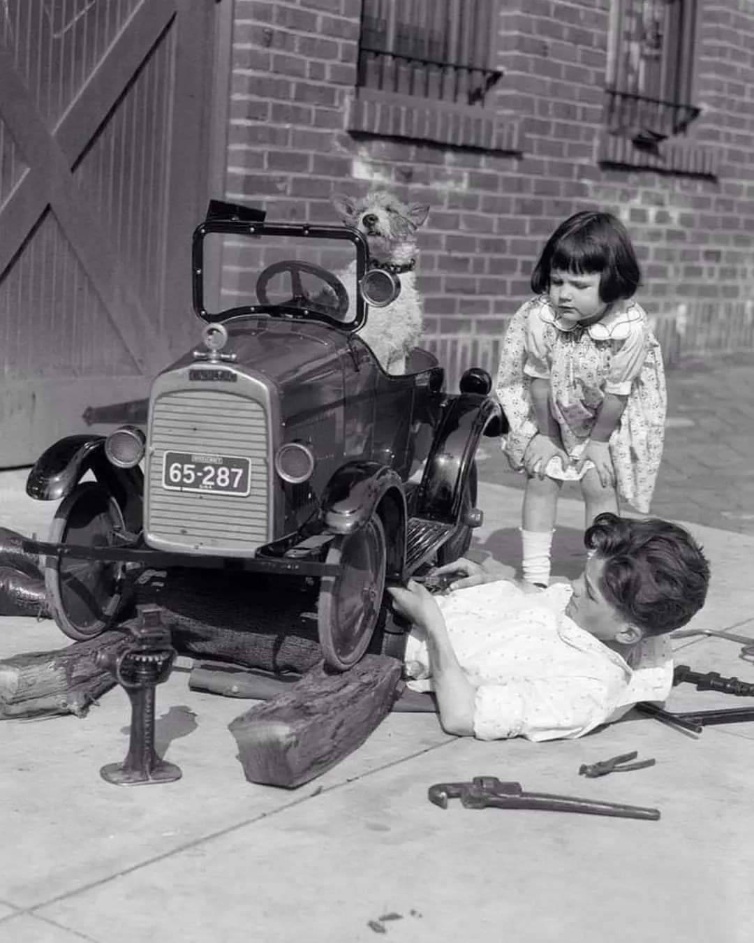 Little girl leaning down watching boy working under toy car with dog behind wheel (1920s).jpg