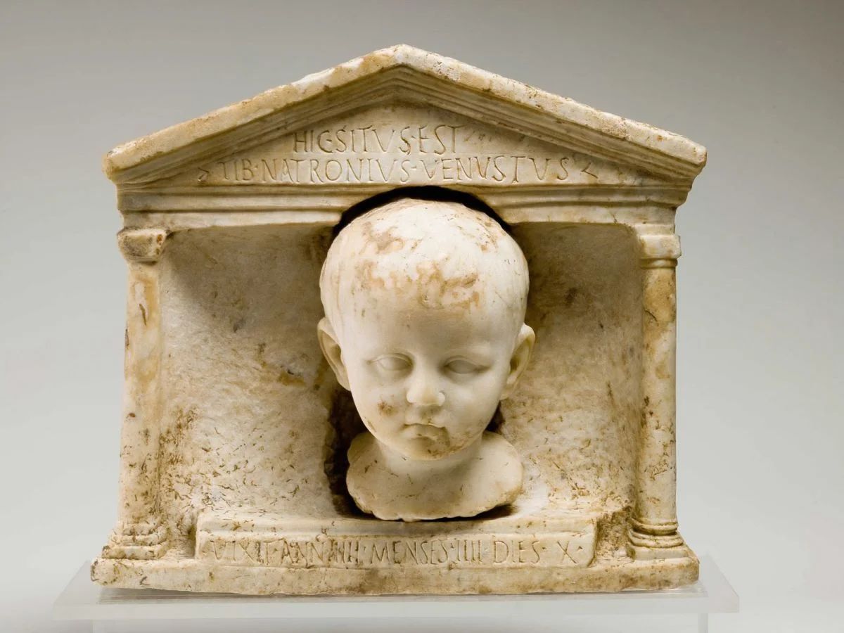 Roman tombstone with the image of a small child’s head. On the stone there is an inscription 'Here lies Tiberius Natronius Venustus. He lived 4 years, 4 months, and 10 days'.jpg