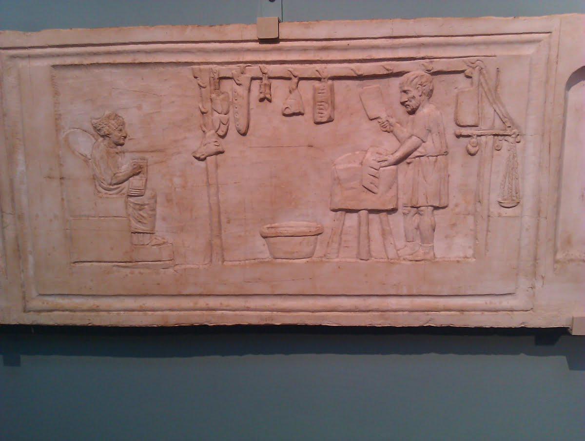 Roman tombstone showing a scene from the work of the deceased, who was a butcher. The man is at work, pieces of meat are hanging in front of him, and the wife is writing something on a wax tablet.jpeg